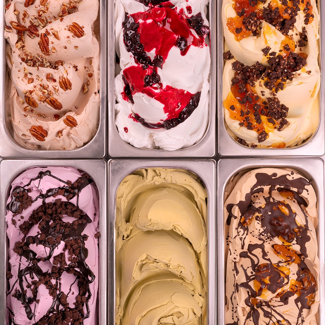 Enam dulang logam gelato dengan pelbagai perisa, termasuk pekan, beri, pistachio dan coklat karamel, dilihat dari atas